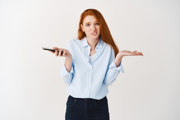 Clueless young woman with red hair and blue shirt shrugging at camera holding smartphone and looking confused dont know white background