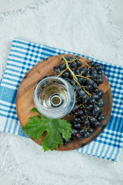 A cluster of black grapes with leaf and a glass of wine on white surface with blue tablecloth