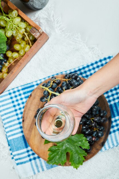 A cluster of black grapes on wooden plate with leaf while hand holding an empty glass