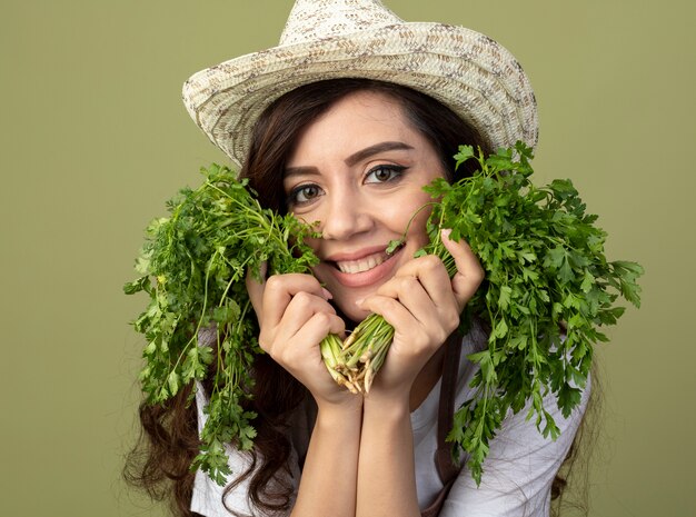 Cmiling young female gardener in uniform wearing gardening hat holds coriander on olive green