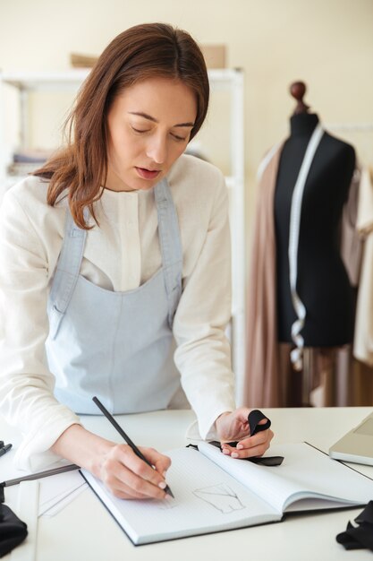 Cocentrated woman seamstress in blue apron drawing new scetch