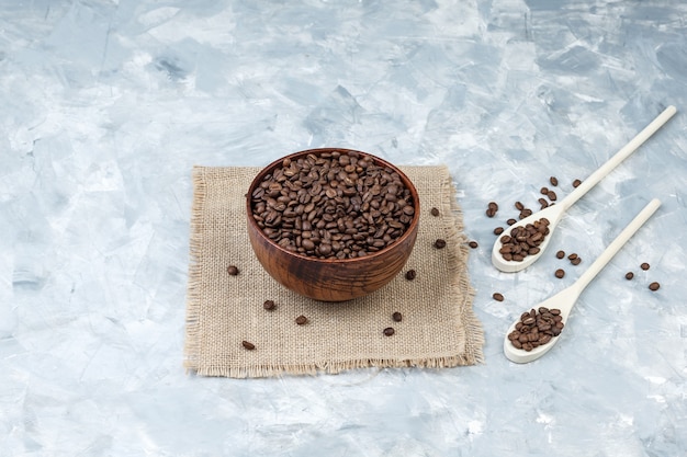 Chicchi di caffè in una ciotola e cucchiai di legno su intonaco e pezzo di sfondo sacco. vista ad alto angolo.