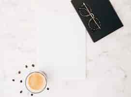 Foto gratuita chicchi di caffè; bicchiere di caffè; carta bianca vuota; occhiali e diario su fondale in marmo