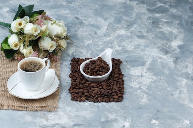 Coffee beans in a white porcelain jug with cup of coffee, piece of sack, flowers high angle view on a blue marble background