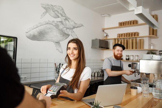Бизнес-концепция кофе Красивая женщина-бариста оказывает платежные услуги клиенту с помощью кредитной карты и улыбается, работая за барной стойкой в современном кафе