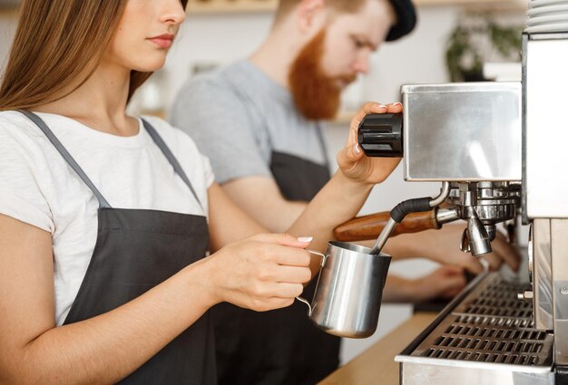 カフェに立っている間彼女のパートナーとのコーヒー注文のためにミルクを準備して蒸しているエプロンの女性バリスタのコーヒービジネスコンセプトの肖像画