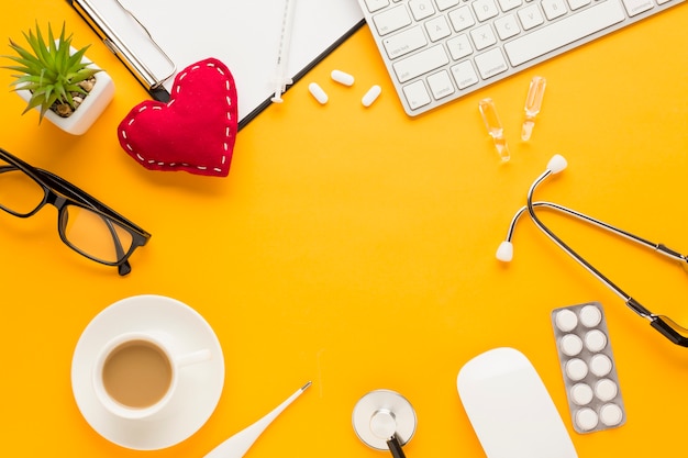 Coffee cup; blister packed medicine; keyboard; eyeglass; succulent plant; thermometer; injection; stitched heart shape; stethoscope; clipboard over yellow backdrop