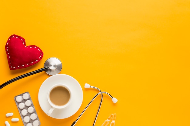 Coffee cup; blister packed tablets; stethoscope and stitched heart shape against yellow background