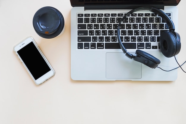 Coffee cup; cellphone and headphone over the laptop against beige background