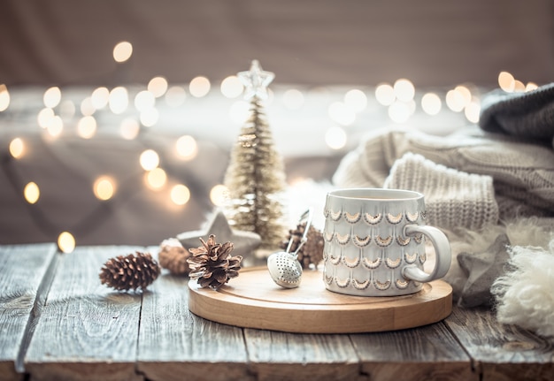 Coffee cup over Christmas lights bokeh in home on wooden table with sweater on a wall and decorations. Holiday decoration, magic Christmas