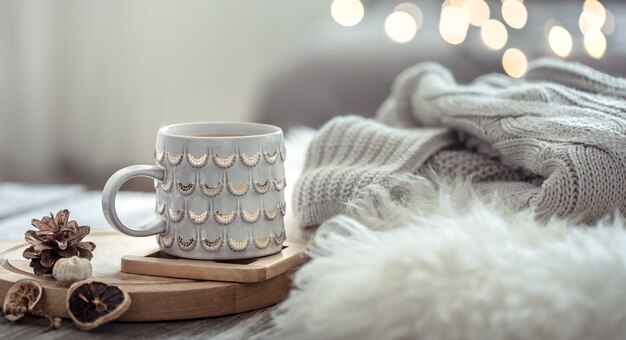 Coffee cup over Christmas lights bokeh in home on wooden table with sweater on a wall and decorations. Holiday decoration, magic Christmas