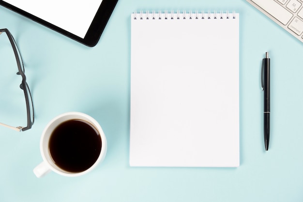 Coffee cup; eyeglasses; digital tablet; keyboard; blank spiral notepad and pen on blue background