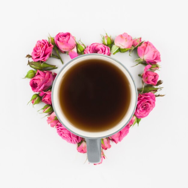 Coffee cup inside the heart shape made with pink roses on white backdrop