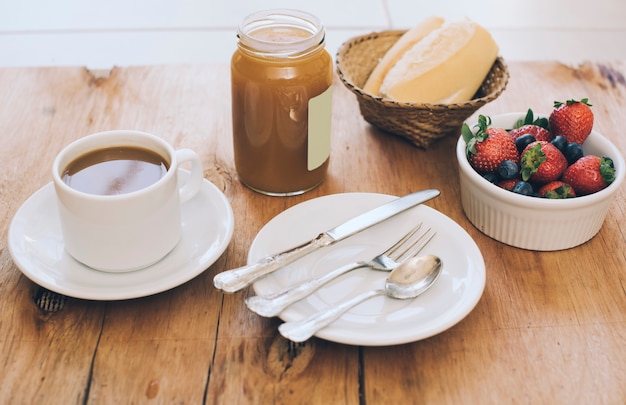 Чашка кофе; набор столовых приборов; баночка с джемом масон; хлеб и ягоды на деревянном столе