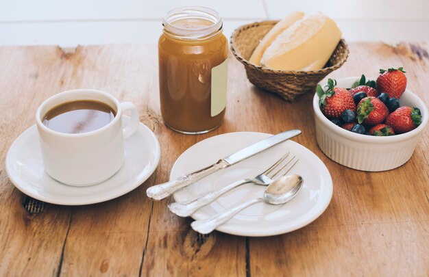 Coffee cup; set of cutlery; jam mason jar; bread and berries on wooden table