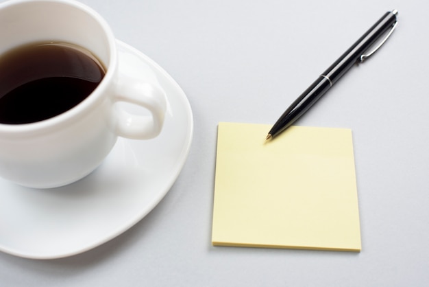 Кофейная чашка с пустой клейкой запиской и ручкой на сером фоне
