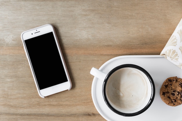 Coffee cup with cookie on white tray and smartphone on wooden backdrop