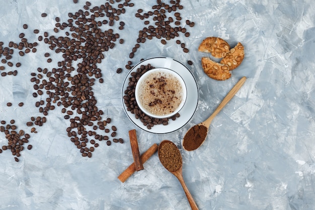 Coffee in a cup with cookies, coffee beans, grinded coffee, cinnamon sticks top view on a grey plaster background