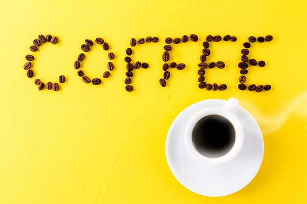 Coffee espresso in small white ceramic cup with coffee beans and word Coffee on yellow vibrant background. Minimalism Food Morning Energy Concept. 