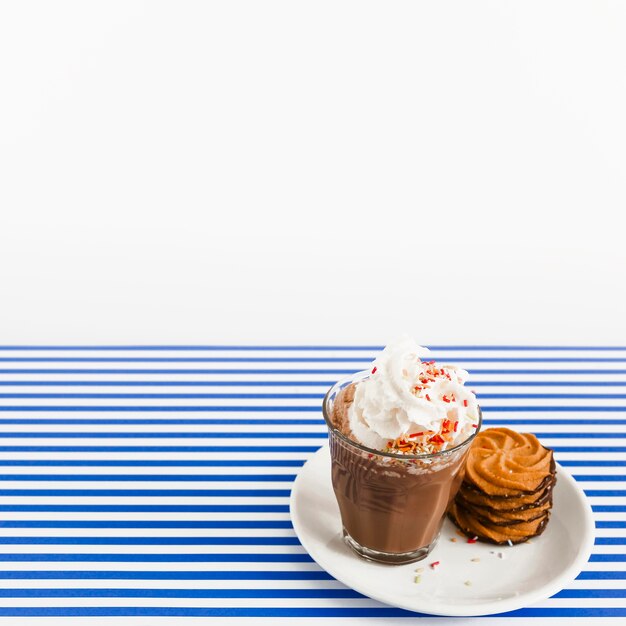 Coffee glass with whipped cream and stack of cookies on plate over backdrop