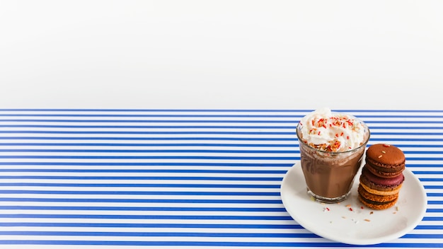 Coffee glass with whipped cream and stack of macaroons on plate over backdrop