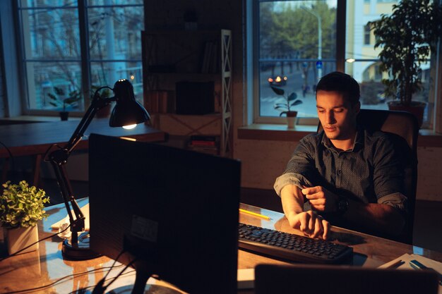 Coffee rescues. Man working in office alone during coronavirus or COVID-19 quarantine, staying to late night. Young businessman, manager doing tasks with smartphone, laptop, tablet in empty workspace.