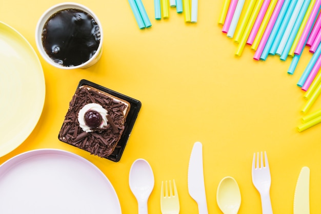 Cold drink; cake slice; empty plate; cutlery and straws on yellow backdrop