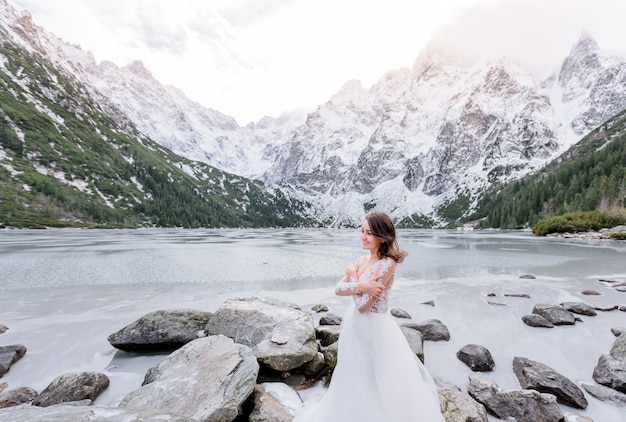 Cold smiled girl dressed in the wedding dress is standing near the frozen highland lake in winter