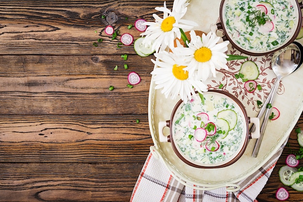 新鮮なキュウリ、木製のテーブルの上にボウルにヨーグルトと大根の冷たいスープ。伝統的なロシア料理-オクローシカ。ベジタリアンの食事。上面図。平置き