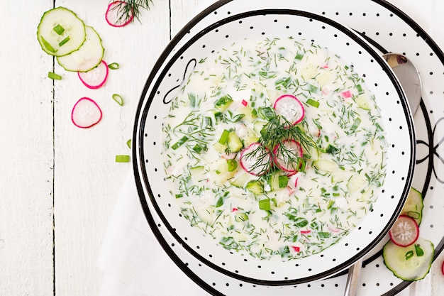 Cold soup with fresh cucumbers, radishes with yoghurt in bowl on wooden table. Traditional russian food - okroshka. Vegetarian meal. Top view. Flat lay