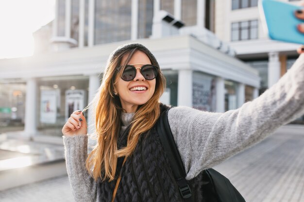 Cold sunny day in city centre of stylish joyful woman making selfie portrait on street. Travelling with backpack, wearing modern sunglasses, woollen sweater, having fun, enjoying leisure.