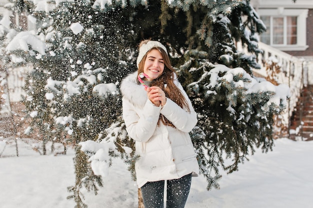 도시의 추운 겨울 날씨와 금요일 나무 배경에 사탕을 들고 재미있고 놀라운 소녀를 즐겼습니다. 따뜻한 옷, 행복한 기분, 내리는 눈, 긍정적인 감정, 새해, 달콤한 얼어붙은 겨울 시간