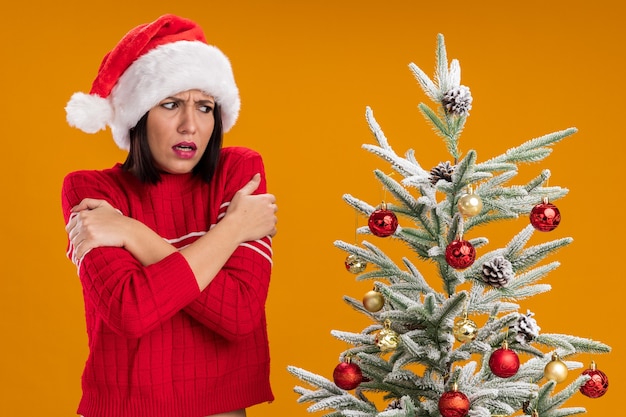 Холодная молодая девушка в шляпе санта-клауса стоит возле украшенной рождественской елки, глядя на нее, скрестив руки на руках, изолированные на оранжевой стене