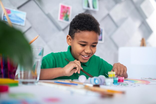 Collapse of pyramid. Cheerful dark-skinned boy in green tshirt is touching destruction pyramid built from tubes of paint for drawing sitting at table against light wall with paintings