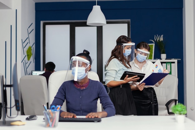 Colleagues doing good work together holding tablet pc weaing face mask for covid19. Multiethnic business team working respecting social distance during global pandemic with coronavirus.