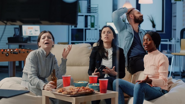 Free photo colleagues having fun with video games and joysticks on tv console after work. workmates enjoying drinks and snacks while playing online game with controllers on television for leisure