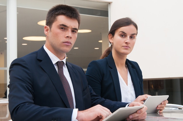 Free photo colleagues looking at camera and holding tablets at desk