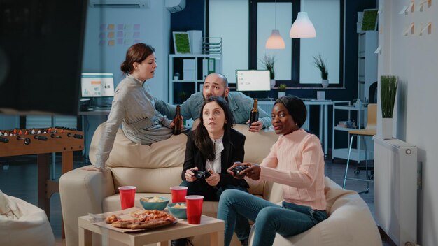 Colleagues playing video games on console with controller after work. Cheerful coworkers enjoying fun activity on free time with drinks and snacks, using joystick on television to play