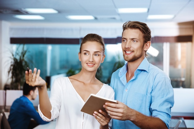 Colleagues smiling, speaking, holding notebook, over office