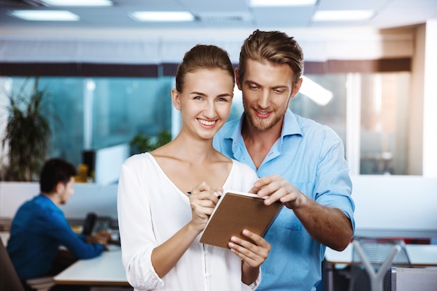 Colleagues smiling, speaking, looking at notebook, over office