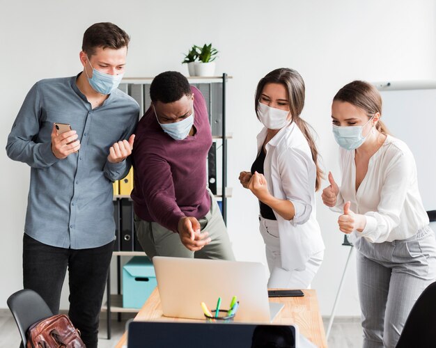 Коллеги на работе в офисе во время пандемии в масках и смотрят на ноутбук