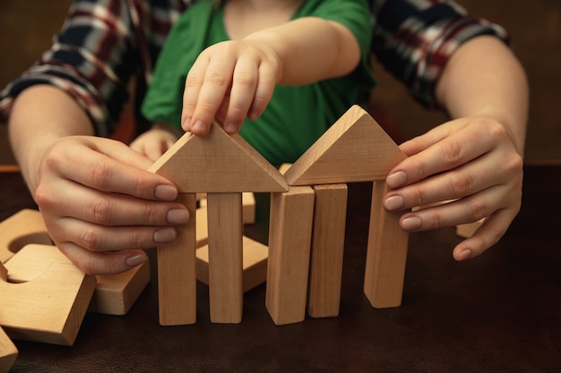 Collecting wooden constructor like house. Close up shot of female and kid's hands doing different things together. Family, home, education, childhood, charity concept. Mother and son or daughter.