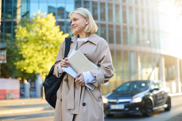 Free photo college lifestyle and student concept smiling blonde woman on street sunny day skyscraper behind her