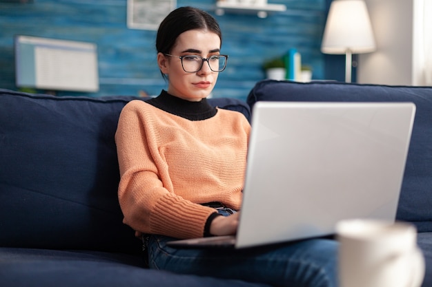 College student with glasses checking email on laptop computer while sitting on sofa in living room. Woman studying commerce information using e-learning university platform