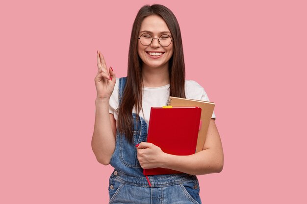 College student with happy look, crosses fingers for good luck on exam, holds notebook for writing records, dressed in denim overalls