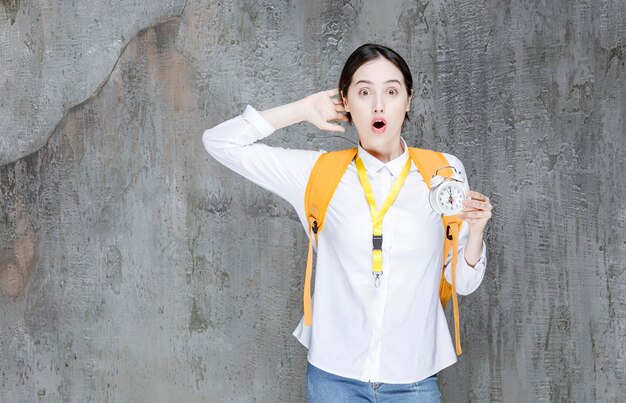 College student with yellow backpack holding clock with surprised expresssion. High quality photo