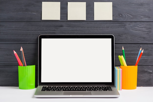 Colored pencils in the plastic holders and laptop on white desk against wooden wall