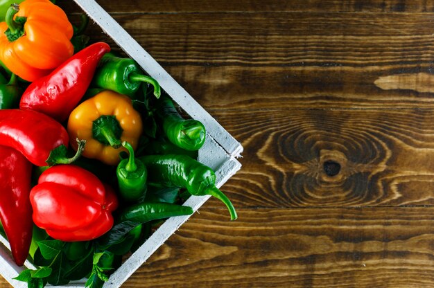 Colored peppers with leaves in a wooden box on wooden surface, flat lay.