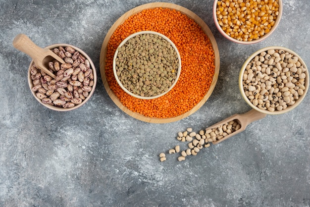 Colorful bowls of various uncooked beans, lentils and corns on marble table surface.