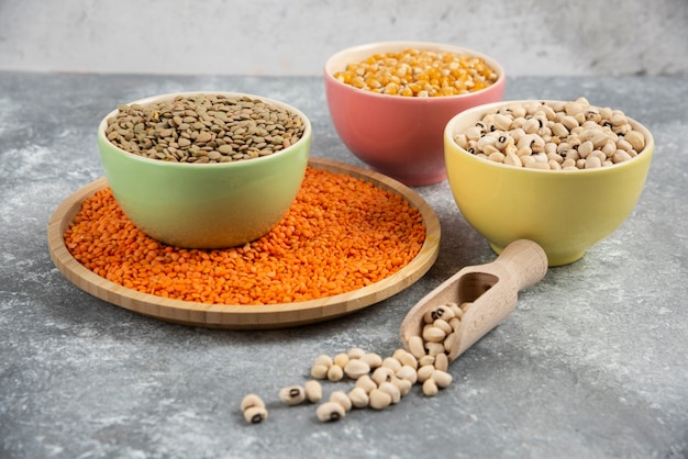 Colorful bowls of various uncooked beans, lentils and corns on marble table surface.
